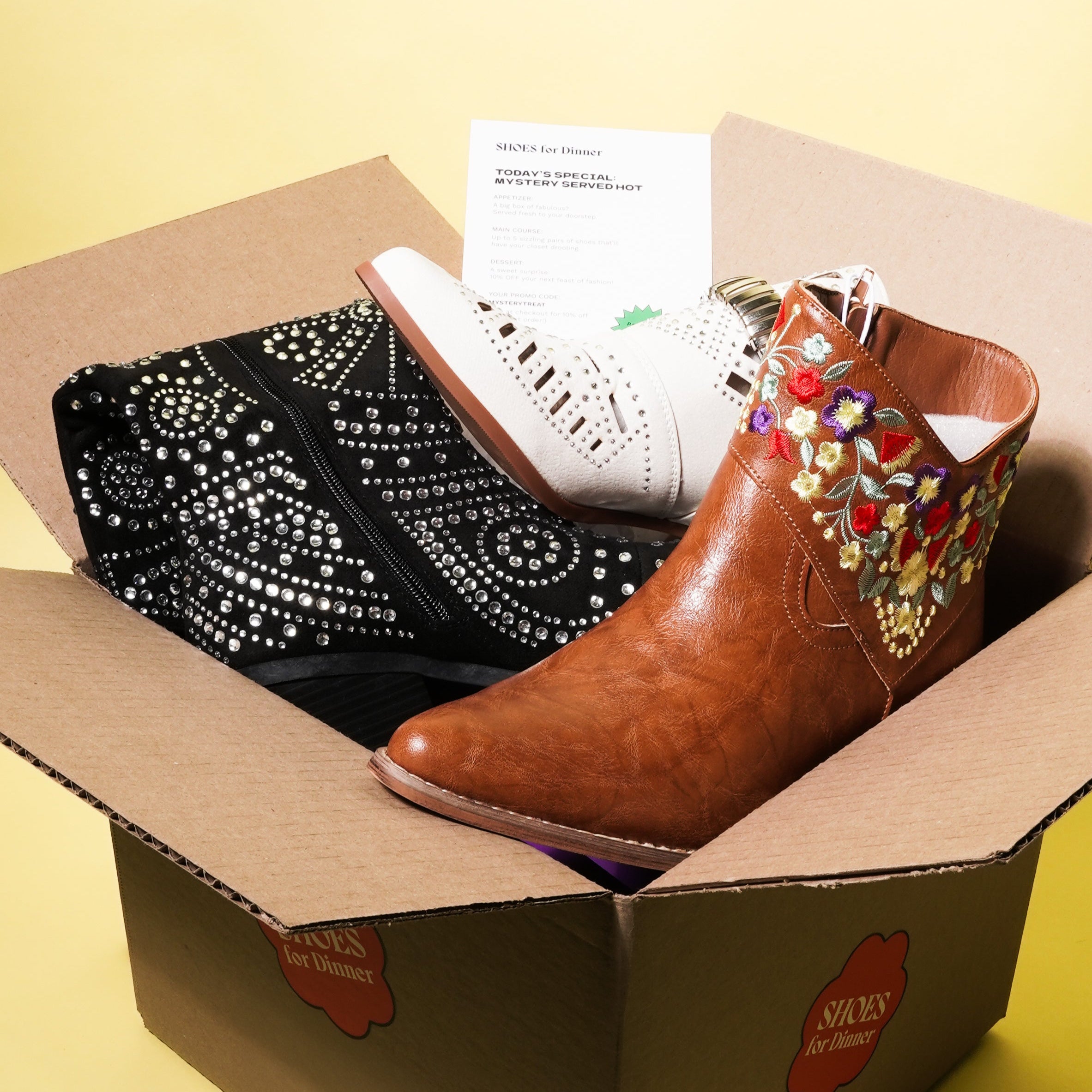 Box containing a black studded boot, white boots, and brown boots with colorful embellishments on a yellow background.