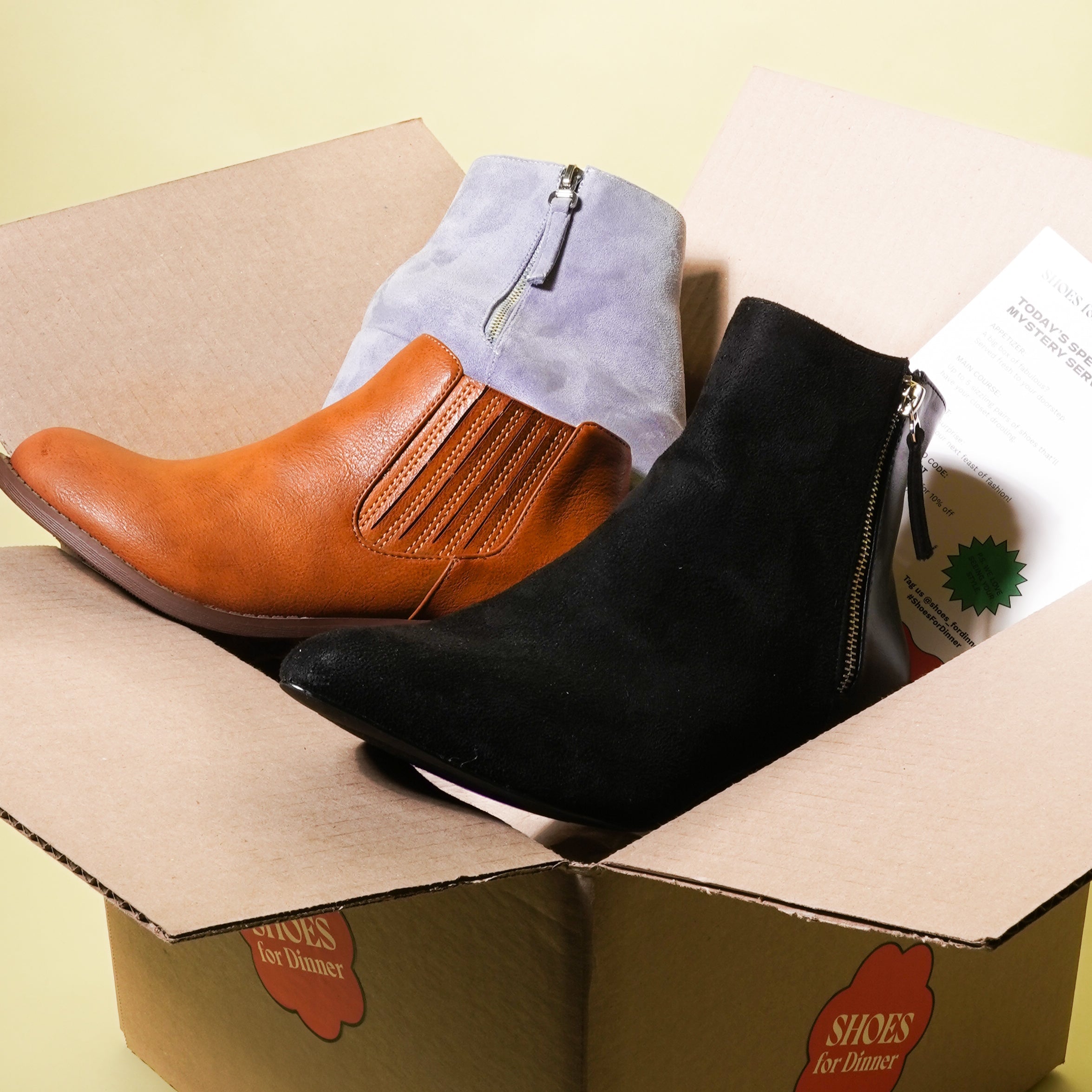 Three pairs of ankle boots in a cardboard box on a light yellow background.